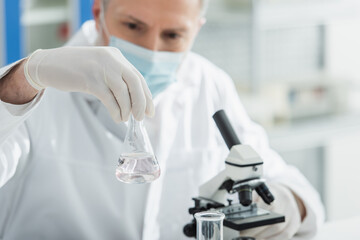 blurred biologist in medical mask holding flask with liquid near microscope.