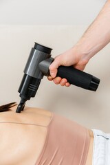 A therapist massages a woman's back with a massage percussion device in her home. The therapist's hand holds a therapeutic vibrating massager. Physical therapy and muscle recovery and massage