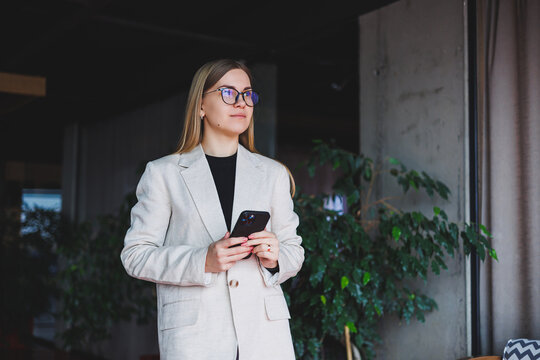 Portrait of a young fair-haired woman in a beige jacket and glasses on her face, a mod phone in the hands of a woman. Beautiful modern blogger girl