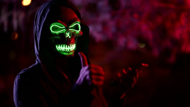 portrait of a person in a terrible glowing mask of death and in a hood against a background of branches illuminated with red light. the character holds the phone, raises thumb and nods. the dark key