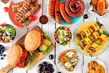 Summer BBQ or picnic food table scene. Assortment of burgers, grilled meat, vegetables, fruits, salad and potatoes. Overhead view on a white wood background.
