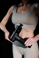 Athletic young woman holding the therapeutic impact massage gun, post-workout recovery. Girl's body in tracksuit against a black background. The nozzles of the vibrating massager. 
