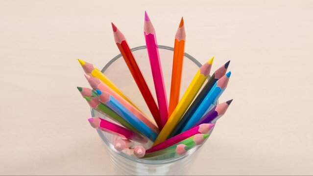 A transparent glass tumbler on the table is filled with colorful wooden pencils. Diversity, variety, rainbow colors. Stop motion animation.
