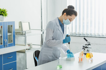 bioengineer in medical mask and white coat working with micropipette and test tubes near microscope.