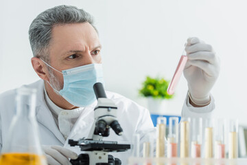 grey-haired bioengineer in medical mask looking at test tube near microscope.
