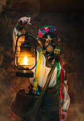 Fototapeta na wymiar Ukrainian woman in a gas mask lights the way with an old gas lamp. Dressed in a traditional folk costume. They hide in the basement, bomb shelters from chemical attacks, with poisonous gases. Vertical