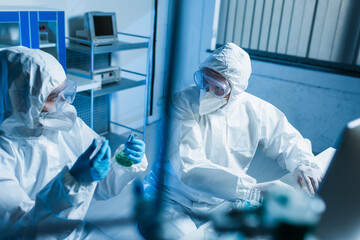 bioengineer with flasks looking at colleague in hazmat suit near computer, blurred foreground.