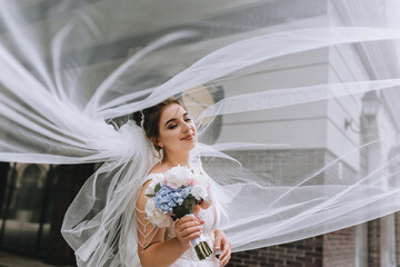 A young, smiling bride with a bouquet in her hands is standing under a long veil. Wedding portrait,...
