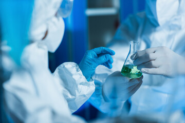 cropped view of bioengineer in latex gloves holding flask with liquid near blurred scientist.
