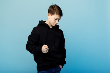 A brooding boy in a black hoodie and jeans is angry on a plain blue background. There is a place for the inscription. Transitional age, crisis of transitional age.