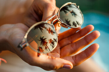 Sunglasses in hands close photo, palm tree reflection and sunset on tropical island, summer mood