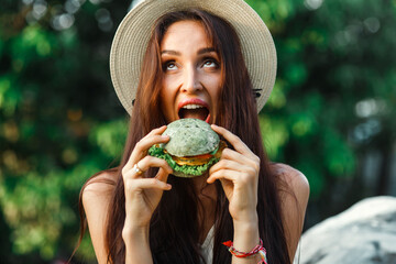 Vegan girl, healthy appearance, holds in her hands and eats a natural, meat-free burger, colored bun, tofu, soy meat, chickpeas. Tropical island Thailand, Bali