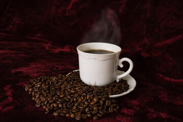 White mug with steaming coffee and beans on red velvet background. 