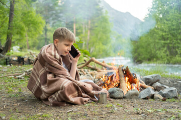 happy teenage boy using phone sitting near fire on camping on mountain