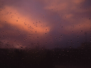 Rain drops on wet glass window with beautiful colourful sunset on background violet sky city roofs below, rainy weather concept 