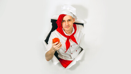 Young handsome man dressed as chef with red tomato. Male cook with vegetable sticking out of hole of white background.