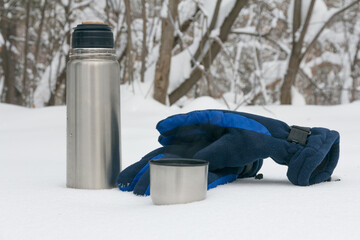 Gloves and a thermos with hot tea against the background of white snow in the forest. Break for hot tea in the forest
