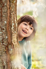 Outdoor portrait of smiling happy toddler boy playing hide and seek hiding behind the tree in spring garden
