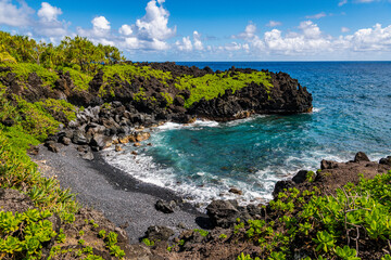 Tropical seascape of a rocky cove with clear blue water and a black sand beach beneath palm trees and lava cliffs at Waiʻanapanapa State Park, Maui - Powered by Adobe