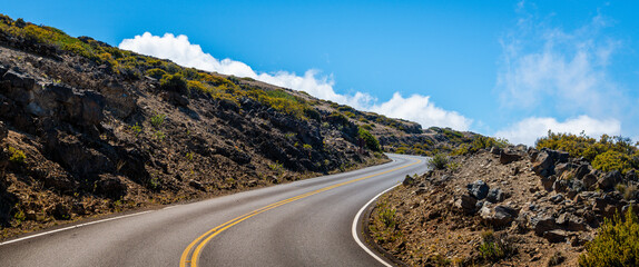 Panorama of mountain road curving through a rocky landscape in the clouds under a blue sky,...
