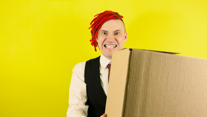 Young crazy man with cardboard box on yellow background in studio. Male courier with bizarre facial...