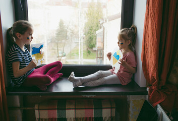 Two little Ukrainian girls with a sad face are sitting on the windowsill, holding a burnt map of...