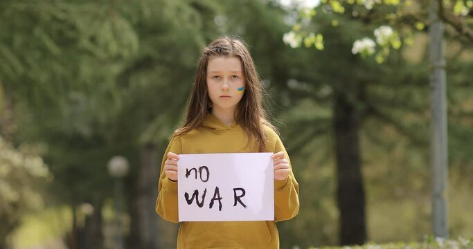 Upset Ukrainian poor toddler girl kid homeless protesting war conflict raises banner with inscription massage text No War outdoors. Crisis, peace, stop aggression, child against Russian war