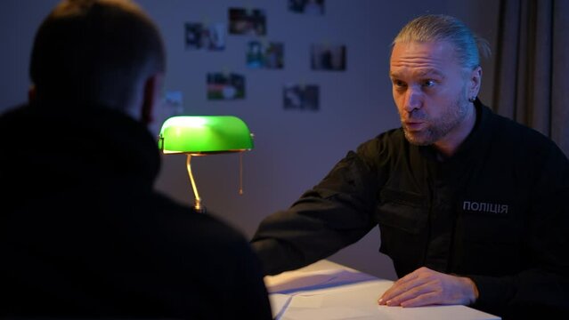 Confident angry policeman talking with suspect passing paper for frank confession. Portrait of serious Caucasian man in Ukrainian uniform interrogating burglar killer indoors. Crime and punishment