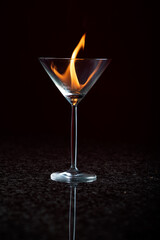 A Single Martini glass on a reflective surface with a black background with a yellow orange flame held within the clear glass