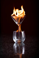 A Martini Glass in a Cognac glass both burning with flowing yellow and orange flames on a reflective surface on a black background