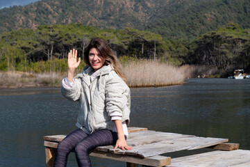 Happy young woman sitting on old small dock. She saying hi with showing her hands. Beautiful landscape view. Nature admiration. Dangling legs from jetty.  