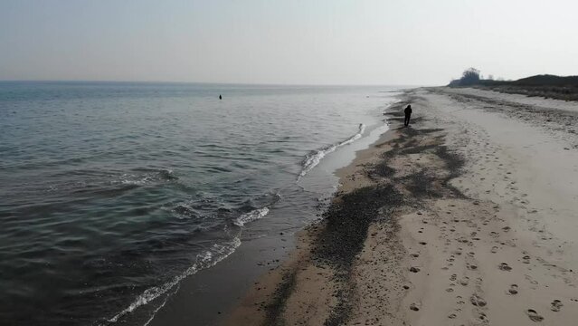 Luftaufnahme von Angler mit Angel in Wathose im Wasser bei Sonne watend per Drohne gefilmt als Fahrt &uuml;ber Strand

