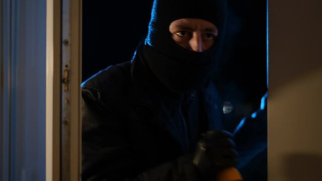 Medium shot portrait of Caucasian thief with flashlight looking inside open house entering leaving. Focused male robber breaking in home at night. Danger and crime concept