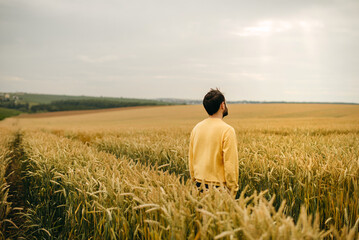 The beautiful bearded man in yellow sweater walking in big field of wheat. Farming, business, young people. Alone, ripe, food, bread. Prayer, self-search, god, rays of sun.  