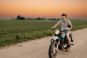 young joyful man rides a retro motorcycle on a country road. The concept of a biker in his youth in the countryside.