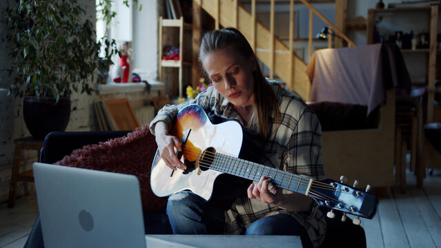 A woman tries to take chords on the guitar and play notes. She is watching videos on her laptop