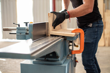 hands of a young carpenter working on an Electric Benchtop Jointer machine cutting a wooden board in a carpentry workshop using safety black gloves