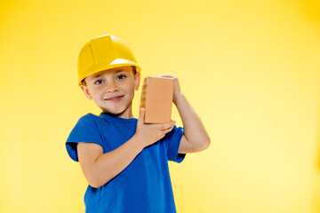 Boy in a construction helmet holds a brick in his hands on yellow background