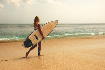 blonde surfing girl walking along a sandy beach with a dock for surfing on the background of a tropical wave - obrazy, fototapety, plakaty