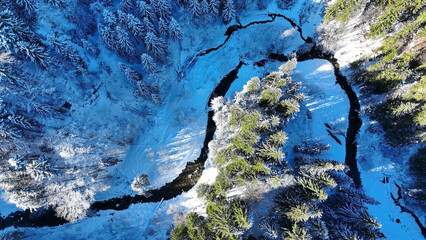 Luftaufnahme mit einer Drohne von einem verschneiten Wald mit Bach im Winter
