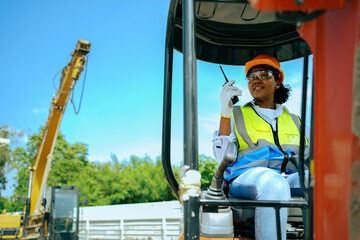 A woman worker driving a backhoe to dig a hole in a construction site holding a walkie talkie.African American female engineer wearing a hard hat and vest.cute female with black skin gender equality.