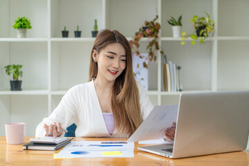 Asian female accountant or banker calculating savings, finance, and economy concepts via laptop. Businesswoman writing charts and graphs that show results in computational analysis.