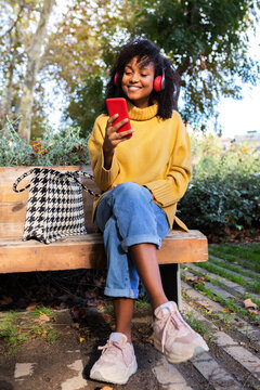 African American woman sit on park bench wearing headphones using mobile phone. Vertical image.