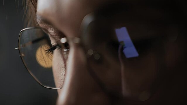 Woman is using phone. Macro shot of female eyes, phone screen is reflected in glasses. User scrolls news or chat in smartphone
