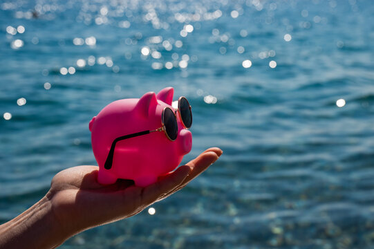 A woman holds a piggy bank in sunglasses on the background of the sea. Budget vacation.