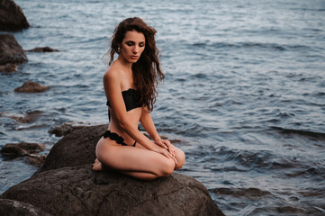 Fototapeta na wymiar Sexy young girl in a black swimsuit sitting on the rocks by the sea in summer on vacation