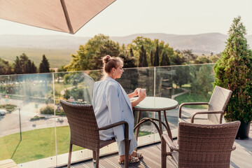 A middle-aged woman sits in a street cafe overlooking the mountains at sunset. She is dressed in a blue jacket and drinks coffee admiring the nature. Travel and vacation concept.