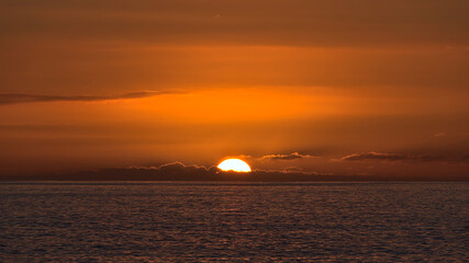View of sunset with sun going down on the horizon between low clouds with dramatic orange colored...