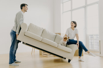 Funny family couple furnish their first home, carry big white sofa, woman teases domestic animal,...