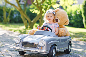 Little adorable toddler girl driving big vintage toy car and having fun with playing with plush toy...